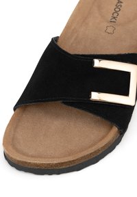 Black suede sandal with a cross strap design, featuring a gold-tone buckle accent and a textured brown insole. Rubber outsole.
