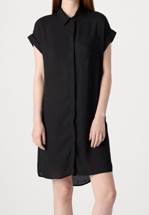 Frau trägt ein schwarzes Hemdblusenkleid mit kurzen Ärmeln, Kragen, Brusttasche und Knöpfen vorne, steht vor einem schlichten Hintergrund.