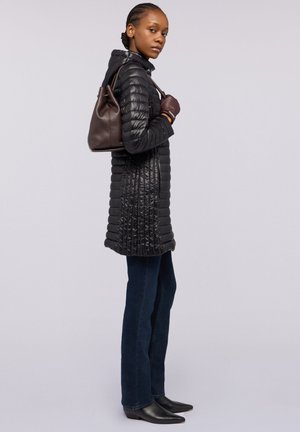 Manteau matelassé noir avec un design texturé, associé à un jean bleu foncé et des bottes noires à bouts pointus. Un sac seau en cuir marron est porté.