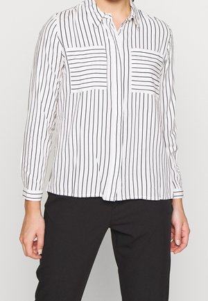 Osoba ubrana w białą koszulę zapinaną na guziki z pionowymi czarnymi paskami oraz czarne spodnie, stojąca na tle jednolitego jasnego tła.