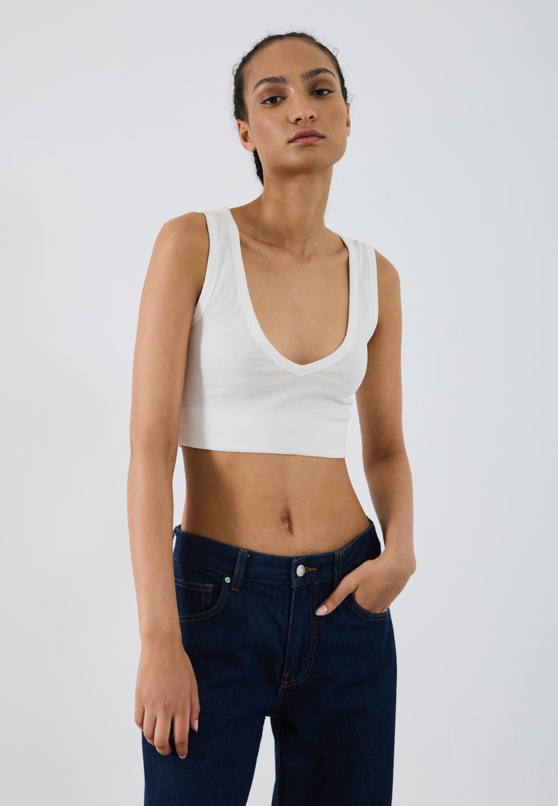 Top alb cu nervuri, scurt, cu bretele late și decolteu adânc în V, asortat cu blugi de un albastru închis, care au cinci buzunare și închidere cu button.