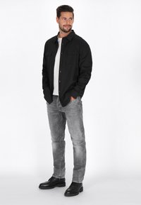Camicia nera con bottoni, dotata di due tasche frontali, indossata sopra una t-shirt bianca. Abbinata a jeans grigi e scarpe nere stringate.