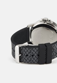 Schwarzes Silikon-Uhrenarmband mit strukturierter Oberfläche und wiederholtem Logo-Muster; silberne Metallschließe und verstellbarer Verschluss am Ende sichtbar.