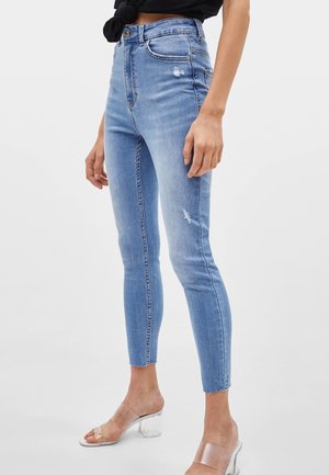 Femme portant un jean skinny taille haute bleu clair avec une légère usure et des sandales à talons transparents, debout devant un fond uni.