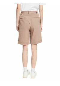 Pantalones cortos beige a medida con dobladillo, dos bolsillos traseros y parte delantera plana. Llevados con zapatillas blancas y calcetines de longitud media.