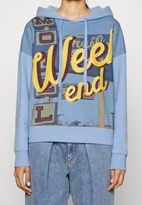 Jasnoniebieska bluza z kapturem z dużą żółtą i bordową grafiką z napisem "Weekend" oraz wzorem znaku motelu. Miękki materiał.