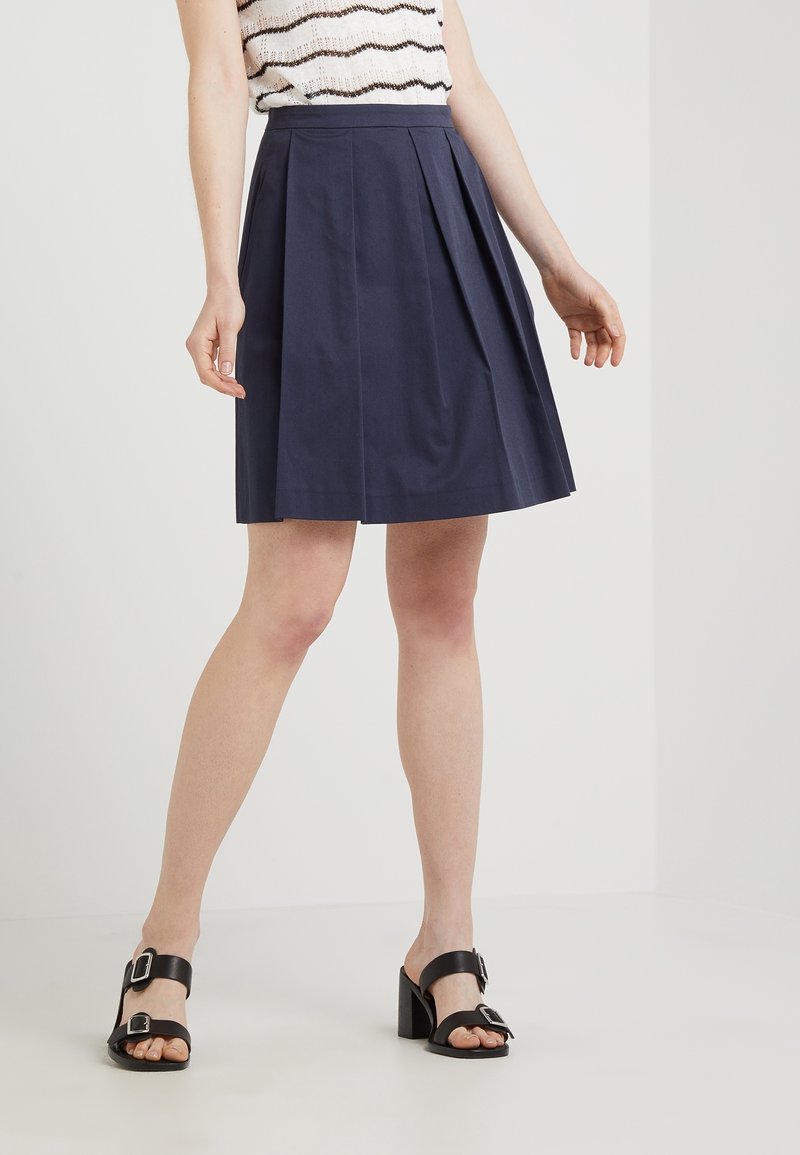 Femme portant une jupe plissée bleu marine longueur genou, un haut sans manches à rayures, et des sandales noires à talons avec des boucles.