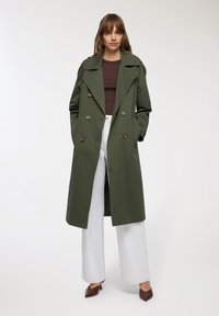 Oliwkowy trencz z podwójnym zapięciem, dużymi guzikami i wyraźnym kołnierzem, noszony na brązowej bluzce i białych szerokich spodniach.