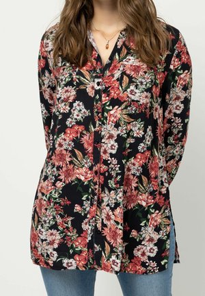 Femme portant un chemisier noir à manches longues avec un imprimé floral rouge, blanc et vert, et un jean bleu, debout les mains derrière le dos.