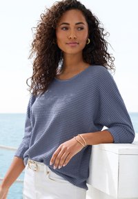 Waffelstrickpullover in Schieferblau mit weitem Ausschnitt und Dreiviertelärmeln, kombiniert mit einem weißen Gürtel und hellen Hosen.
