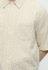 Μπεζ κοντομάνικο πουκάμισο με διακριτικό λεοπάρ σχέδιο και μπροστινή τσέπη στο στήθος, που φοριέται από άτομο με μερικώς ορατό χέρι.