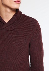 Pull bordeaux en maille avec col châle, texture côtelée et coupe ajustée. Le tissu semble doux et légèrement extensible.