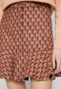 Jupe marron à motifs géométriques avec petits nœuds, portée avec un haut à manches longues couleur pêche clair, main reposant le long du corps.
