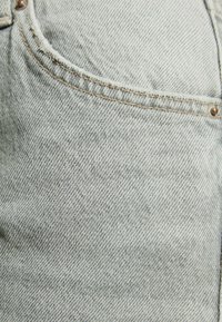 Tela de mezclilla gris claro con una textura sutil, que presenta costuras en contraste y un bolsillo visible con un acento de remache metálico.
