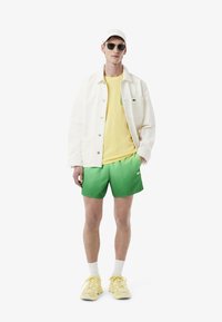 Veste en denim blanche, t-shirt jaune, short dégradé vert, baskets jaunes, casquette blanche, lunettes de soleil grises. Design simple et sportif.