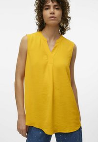 Top giallo senza maniche con scollatura a V, tessuto testurizzato e vestibilità comoda. L'orlo è leggermente curvato, abbinato a pantaloni in denim con decorazioni.