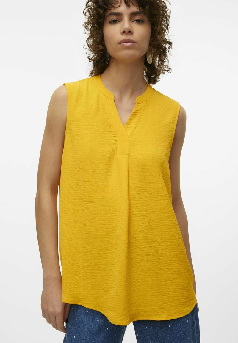 Top giallo senza maniche con scollatura a V, tessuto testurizzato e vestibilità comoda. L'orlo è leggermente curvato, abbinato a pantaloni in denim con decorazioni.