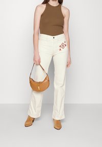 Kvinna som bär en brun ärmlös topp, vita jeans med röd broderi på fickan, tan mockastövlar och håller i en liten brun handväska.