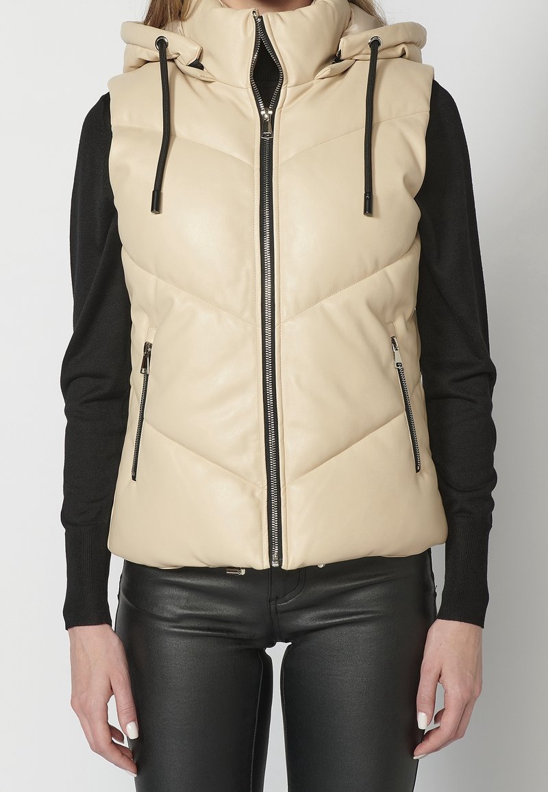 Veste sans manches matelassée beige avec fermeture éclair devant et capuche, portée sur un haut noir à manches longues et un pantalon en cuir noir.