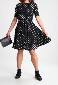 Czarna sukienka w kropki z krótkimi rękawami i dopasowanym gorsetem, rozszerzająca się w talii, w towarzystwie czarnej kopertówki i butów na obcasie.