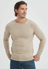 Pull en tricot beige à manches longues, col rond et poignets côtelés. Le tissu a une texture lisse et un motif chiné subtil.