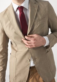 Beige blazer met een gestructureerde weving, één knoop en twee voorzakken. Gecombineerd met een wit overhemd en een rode stropdas.
