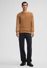 Mann steht in einem strukturierten braunen Pullover, schwarzen Jeans und schwarzen Schuhen vor einem schlichten hellen Hintergrund.