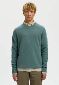 Pull en tricot teal avec un col rond et des poignets côtelés, superposé sur une chemise à col rayée, associé à un pantalon khaki.