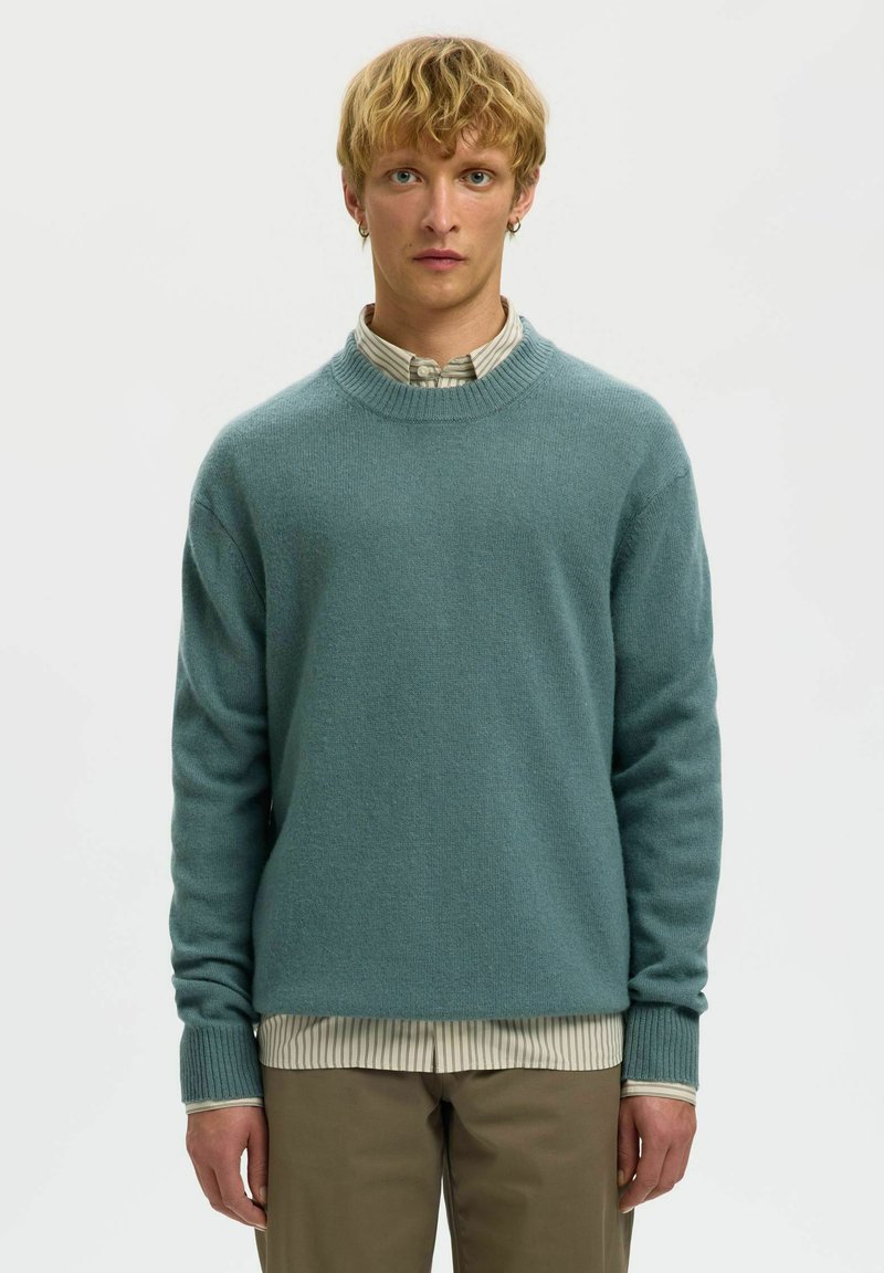 Pull en tricot teal avec un col rond et des poignets côtelés, superposé sur une chemise à col rayée, associé à un pantalon khaki.