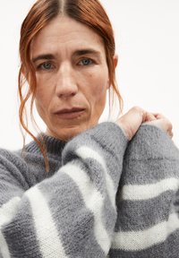 Hellgrauer Strickpullover mit weißen Streifen, der eine weiche Textur und einen breiten, gerippten Ausschnitt aufweist. Die Ärmel sind leicht oversized.