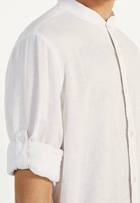 Camicia bianca in lino con colletto alla cinese e maniche arrotolate fissate da una linguetta con bottone sul braccio.