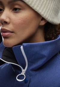 Blauwe, met een rits afgesloten jas met een hoge kraag en witte ritsaccessoires. Draagt een crème kleurige ribbelgebreide beanie. Gladde textuur en gestructureerd ontwerp.