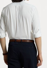 Camisa blanca de manga larga con puños enrollados, combinada con pantalones oscuros y un cinturón de cuero marrón. Costura trasera visible y bolsillos en los pantalones.