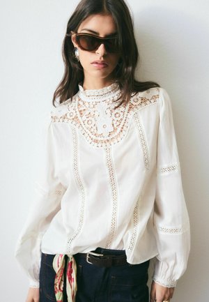 Frau mit dunkler Sonnenbrille, weißer Spitzenbluse mit hohem Kragen, dunkler Jeans und einem bunten Schal, der in der Taille gebunden ist.