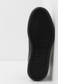 Semelle de sneaker en caoutchouc noir avec des rainures texturées et un marquage en relief ; les côtés lisses se rétrécissent vers le talon pour un design épuré.