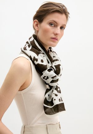 Woman wearing sleeveless ribbed top and black-and-white patterned scarf, looking sideways against plain background.