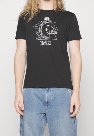 Person iført sort t-shirt med hvidt abstrakt ansigtsdesign, tårdrop, fugle og teksten "Makia", kombineret med lyseblå denimjeans.