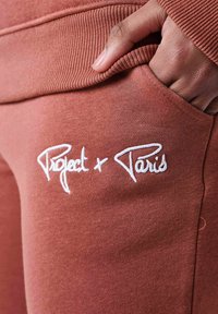 Joggers de color óxido con una tela suave y texturizada. Cuenta con un logotipo bordado en blanco "Project + Paris" en el área del bolsillo izquierdo.