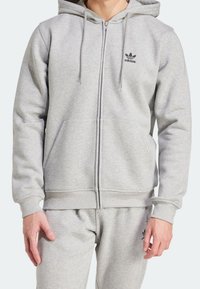 Light grey zip-up hoodie in soft fleece material with a front kangaroo pocket and black Adidas logo on the chest, featuring drawstring hood.