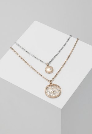 Deux colliers à chaînes superposées, l'un en argent avec un pendentif rond lisse, l'autre en or avec un pendentif rond à motif floral ajouré, présentés sur un bloc blanc incliné.