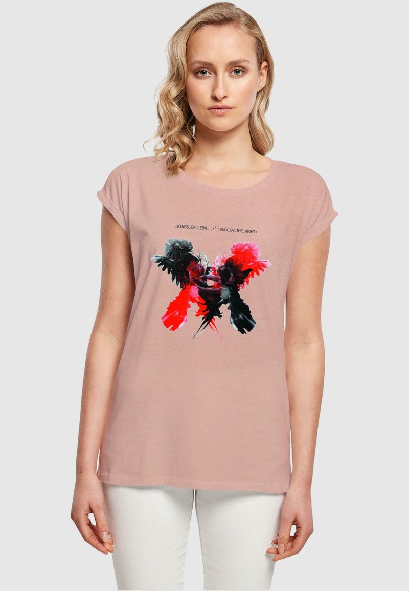 Rosa T-Shirt mit kurzen Ärmeln, das ein grafisches Design mit roten, schwarzen und pinken Elementen sowie Text zeigt, getragen mit hellen Hosen.