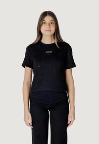 Cropped t-shirt nera in cotone, con applicazioni di borchie scintillanti e il logo "GUESS" in argento sul petto. Indossata con pantaloni neri.