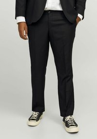 Schwarze taillierte Hose mit geradem Schnitt, ausgestattet mit einem Knopfverschluss; kombiniert mit schwarz-weißen Sneakers. Glatte Stofftextur.