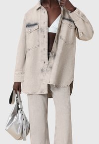 Personne à la peau foncée portant une veste en jean clair délavée et un pantalon assorti, tenant un sac à main argenté métallisé, haut blanc visible sous la veste.
