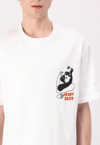 Camiseta de algodón blanca con un gráfico impreso de una figura abstracta con manos y el texto "HEART BASED" en color naranja. Mangas cortas.