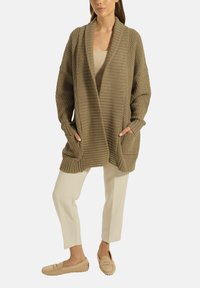 Frau trägt einen langen, strukturierten braunen cardigan mit Taschen, ein beiges Oberteil, knöchellange cremefarbene Hosen und beige Slipper, steht mit den Händen in den Taschen.