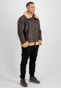 Chaqueta bomber de cuero marrón con cuello y puños de piel sintética beige; cierre con cremallera en el frente, bolsillos laterales y un corte holgado. Pantalones cargo negros y zapatillas deportivas.