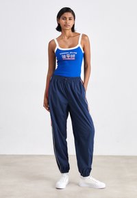 Blå ribbestrikket tanktopp med hvit kant og logo, sammen med marineblå joggebukser med røde striper og elastiske mansjetter. Hvite sneakers fullfører antrekket.