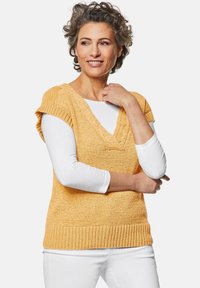 Geel gebreide trui met korte mouwen en een V-hals, gelaagd over een witte lange mouwen shirt. Ribbelboord en textuurafwerking.