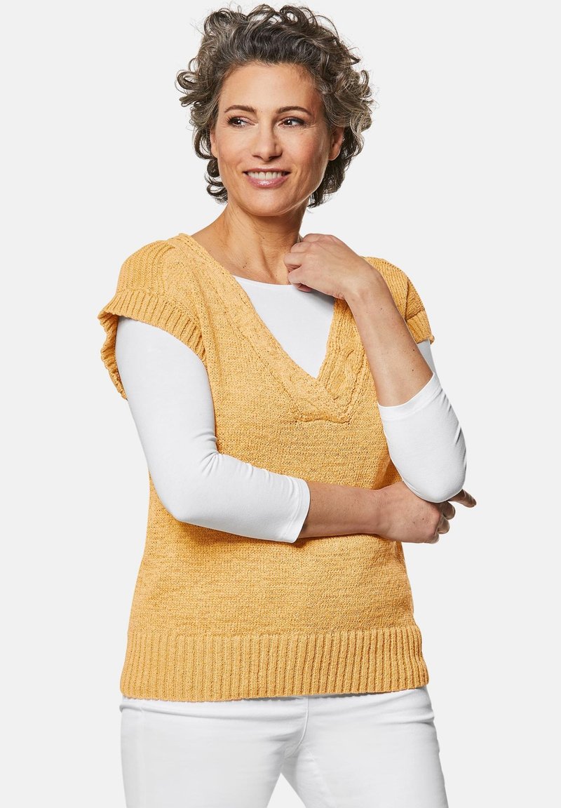 Geel gebreide trui met korte mouwen en een V-hals, gelaagd over een witte lange mouwen shirt. Ribbelboord en textuurafwerking.